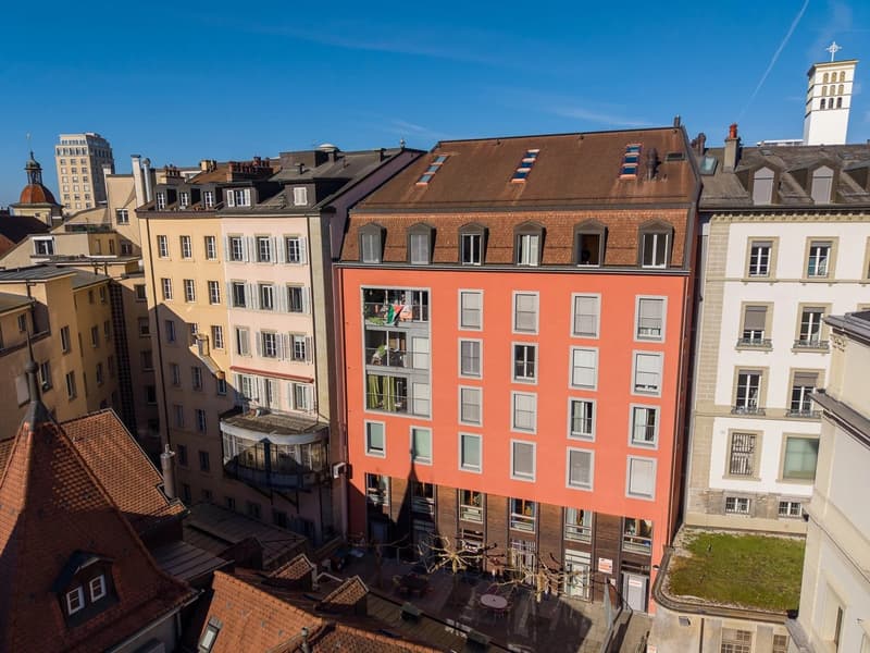 Aerial view of a multi-story building with multiple balconies, red brick facade, brown roof, multiple windows, and a small garden in front.