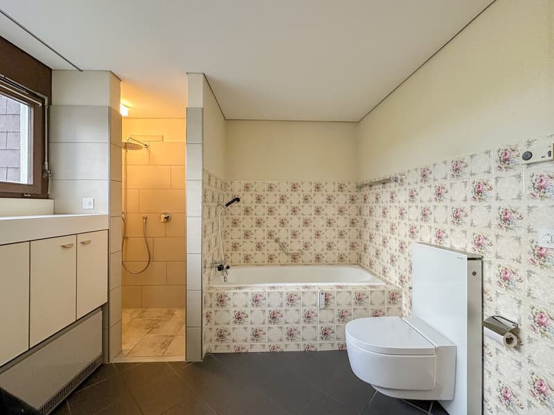 Bathroom with walk-in shower, bathtub, and toilet; floral tiles and white cabinets; window in the shower area