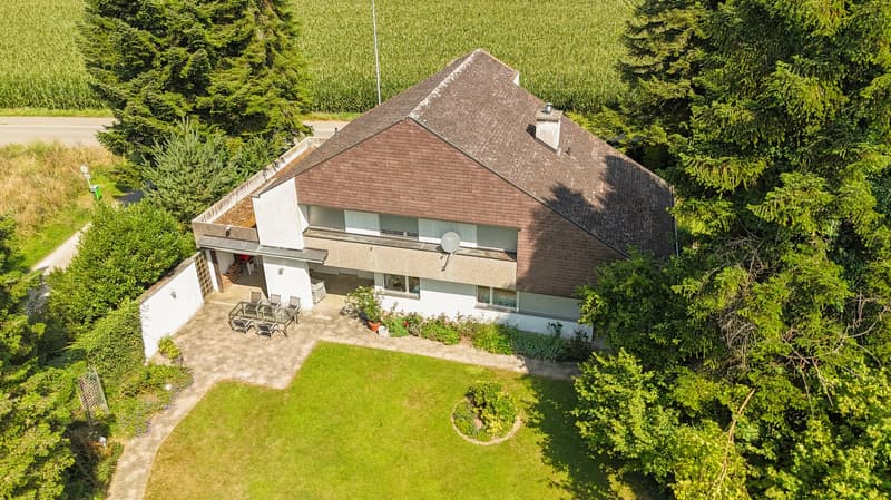 House on raised ground floor, 2 stories, brown roof, white exterior, garden with plants and trees, green lawn, patio with table and chairs, satellite dish, view of the surrounding fields