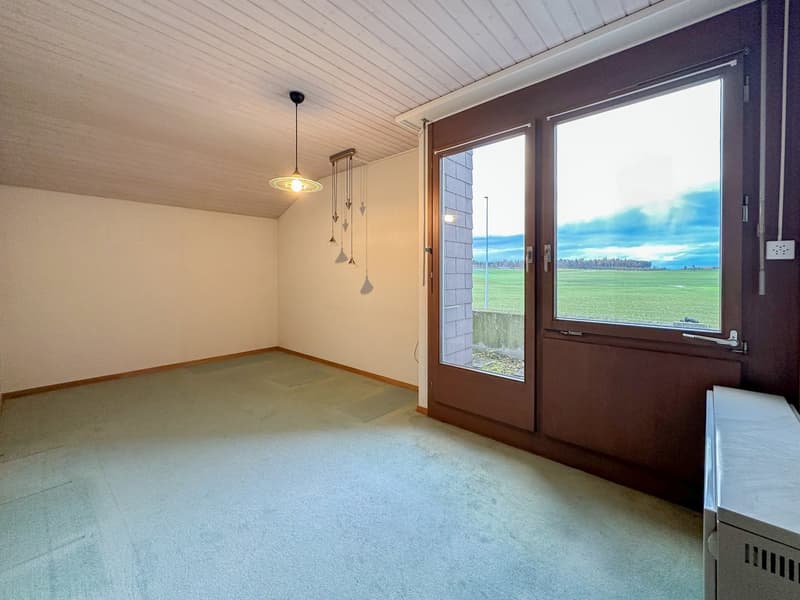 Empty room with large windows, wood-paneled ceiling, beige walls, gray carpet, glass double doors, outside view of a field, hanging lamp