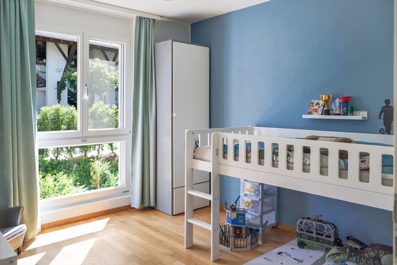 Spacious bedroom with blue walls, large windows overlooking a garden, a white bunk bed, a wardrobe, and shelves displaying toys and other decorative items.