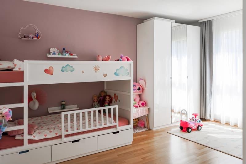 White crib with drawers, white wardrobe, shelves with toys and dolls, pink and white color scheme, hardwood floor, large window