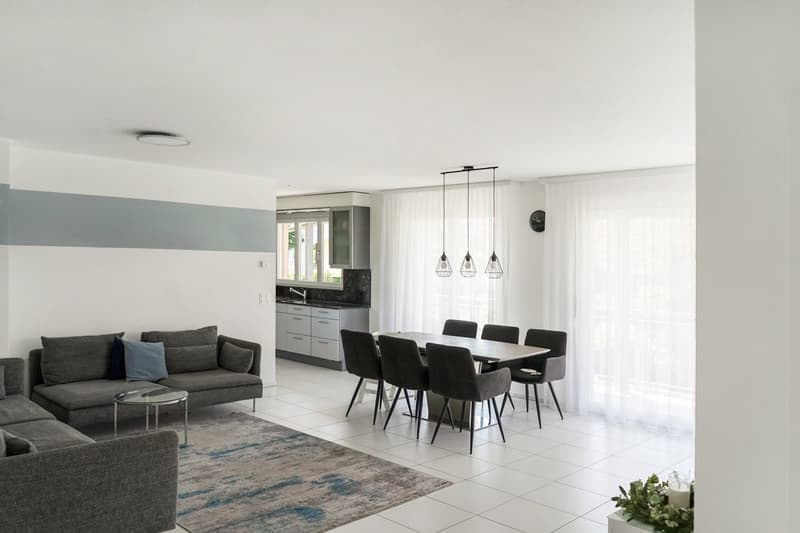 The image shows an open concept living room and dining room with a modern, minimalist design. The living room features a gray sofa and a coffee table, while the dining area has a rectangular dining table with black chairs. The kitchen is visible in the ba