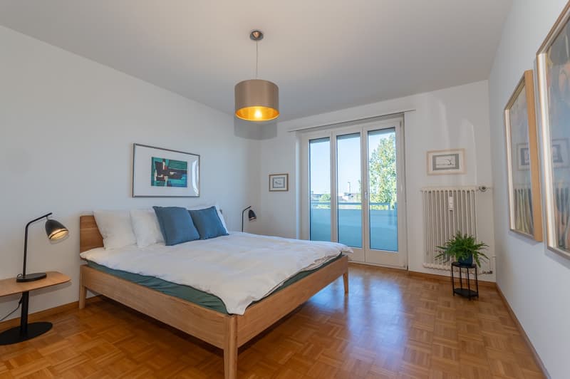 wooden bed, white bedding, blue pillows, lamp, wooden floors, white walls, large window with balcony access, modern artwork