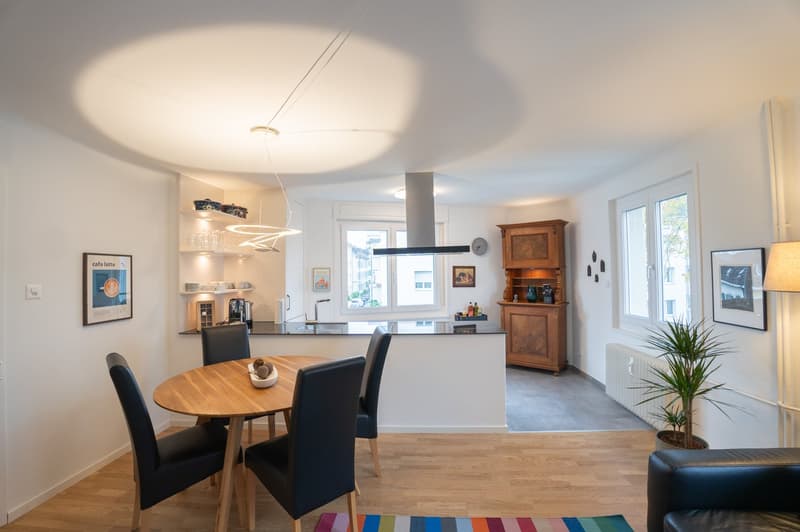 The image shows an open concept living space with a kitchen, dining area, and living room. The kitchen features a black range hood and built-in appliances. The dining area has a round wooden table with black chairs. The living room has a wooden cabinet an