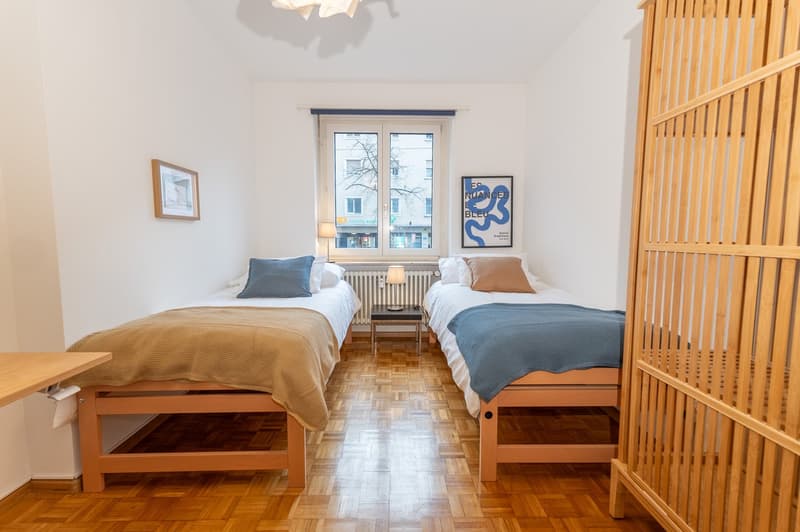 Room with two single beds, nightstands with lamps, white walls, wooden floor, window, radiators