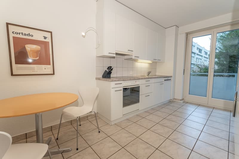 Modern kitchen with white cabinets, white tiled floor, stainless steel appliances, wooden dining table, and white chairs.