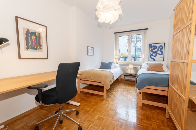 room with two single beds, desk, office chair, wall lamp, night lamp, framed pictures, wooden furniture, radiator