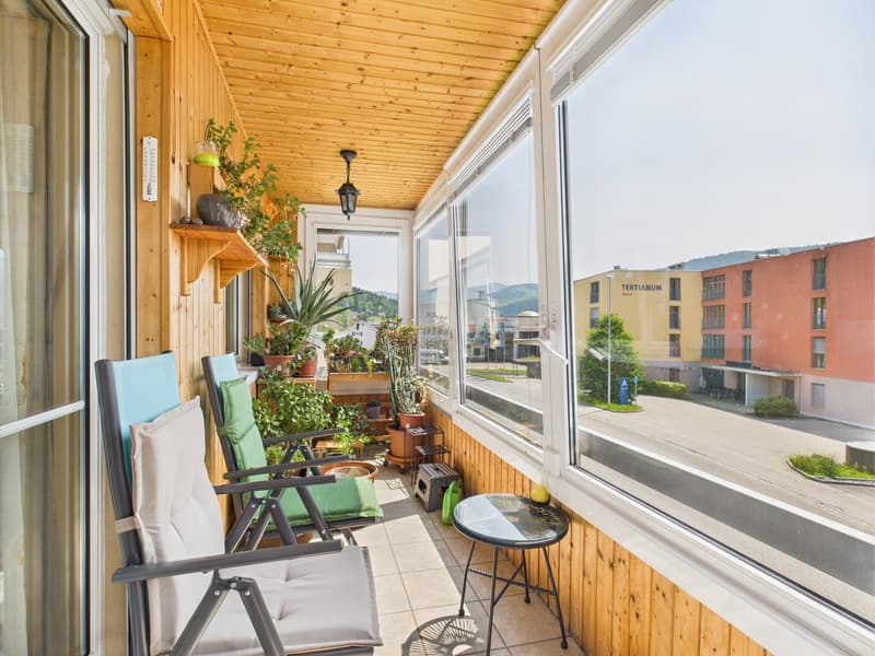 Cozy balcony with wooden ceiling, large windows offering a view of the surrounding buildings, potted plants and flowers, comfortable chairs, and a small table.
