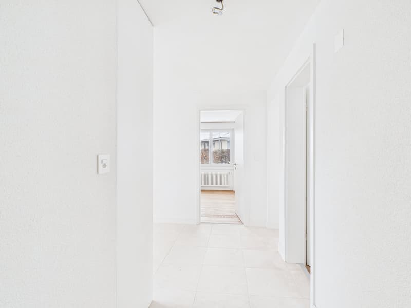 Empty hallway with white tiles, white walls, ceiling light, window, door leading to a room