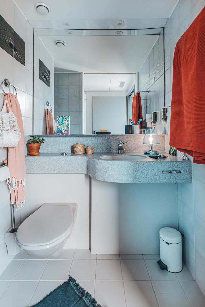 White tiled walls and floor, large mirror, large sink with countertop, white toilet, small trash bin, orange towels on towel rack, a potted plant on countertop, trash paper roll on wall, a rug at the floor
