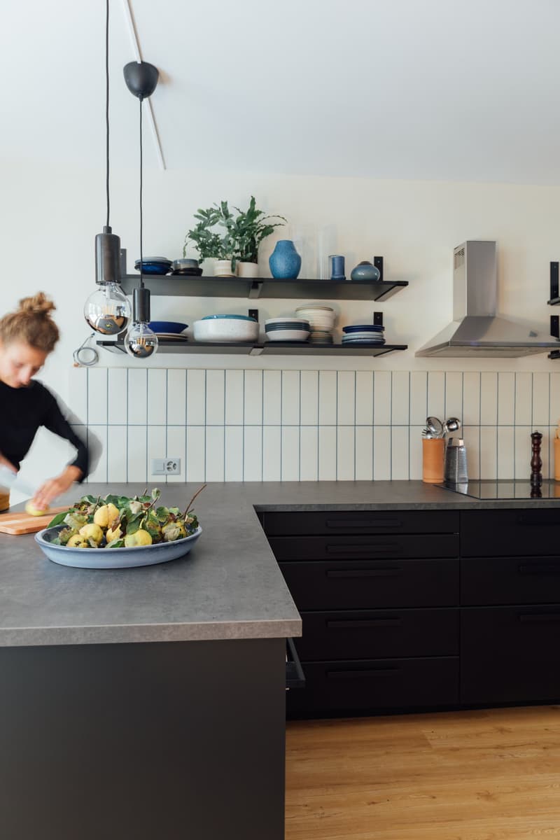modern kitchen with light color tile, gray countertops, black cabinets, 3 hanging lamps, white tiles on wall, plate rack, and stove