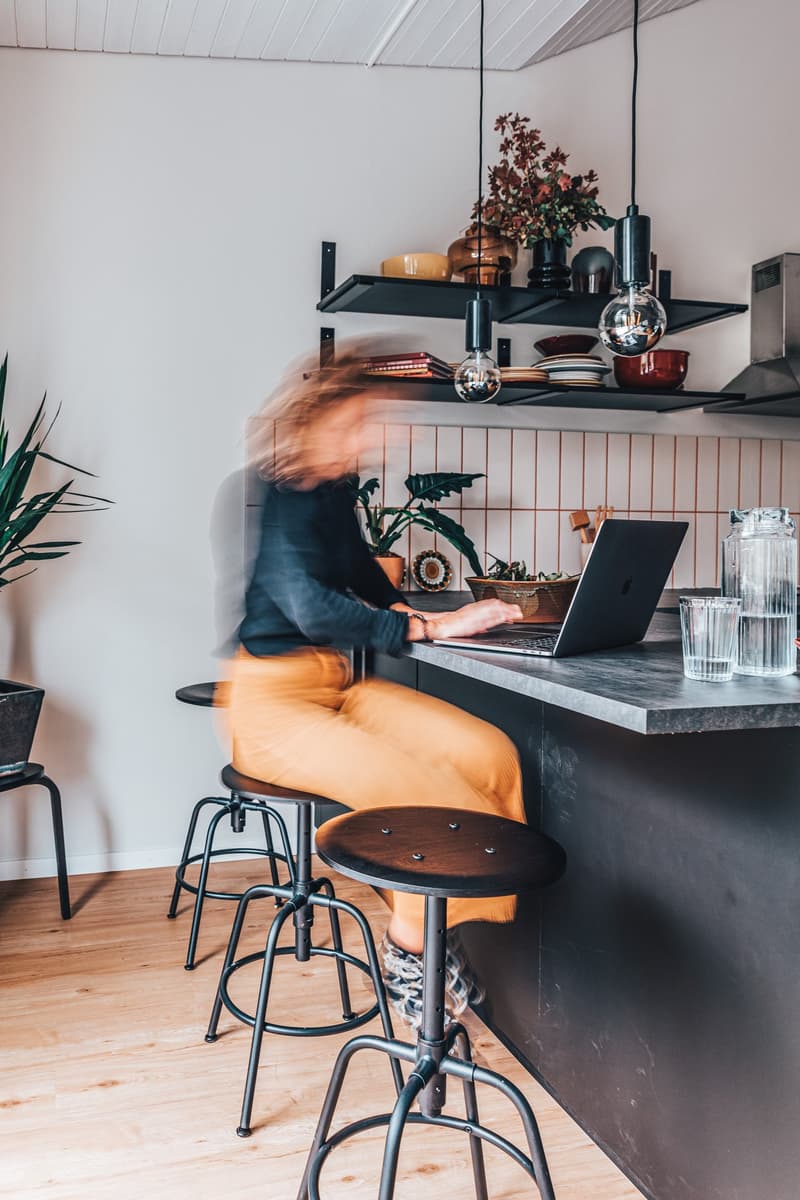 A person working on a laptop at the kitchen bar, with a modern industrial design, wooden floor, and potted plants.