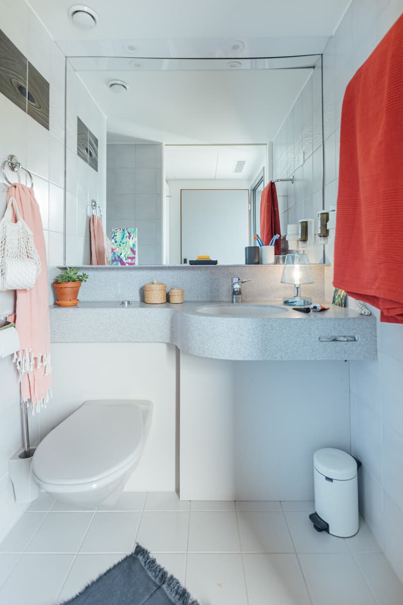 Bathroom: sink, toilet, mirror, small potted plant, trash can, towels, rug, white tiled floor