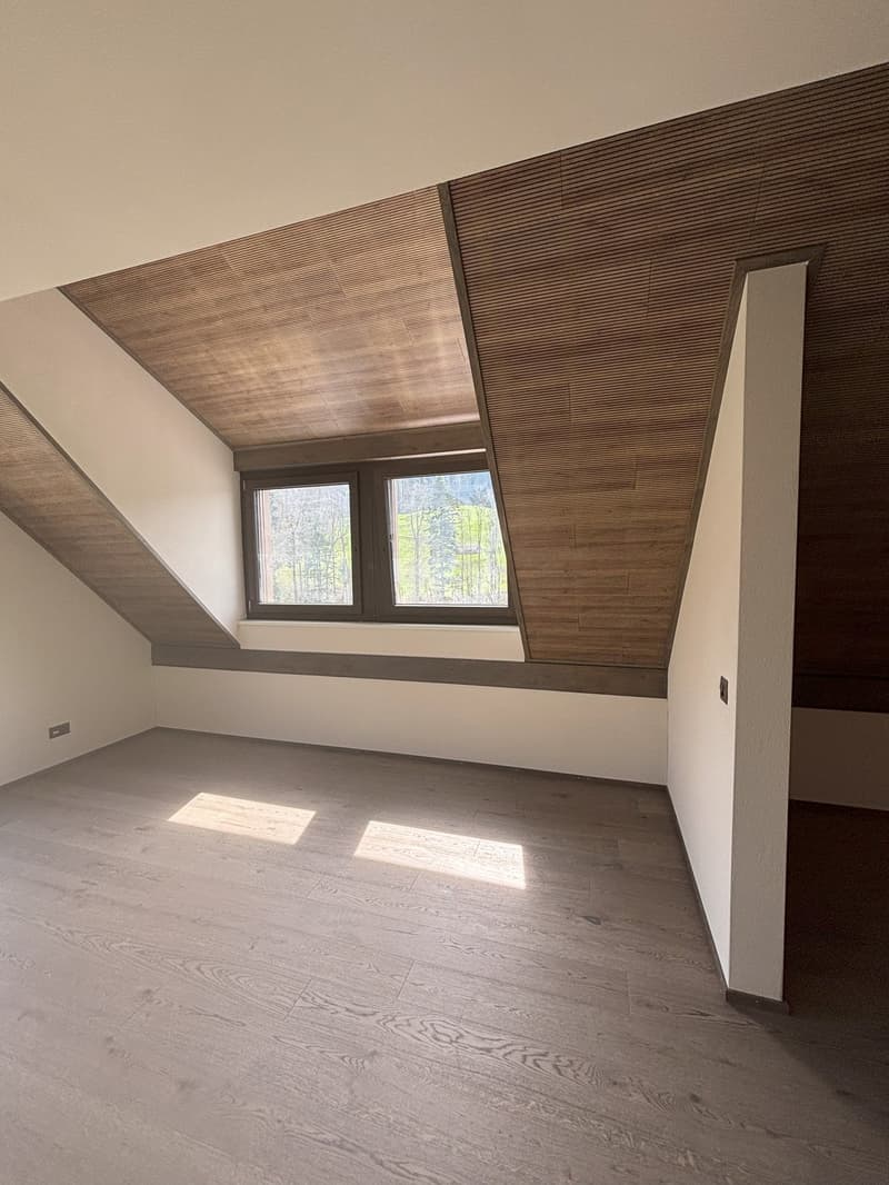 Spacious attic room with slanted wooden ceiling, large windows overlooking the outdoors, and hardwood flooring. The room has a modern, minimalist design with clean lines and neutral colors.