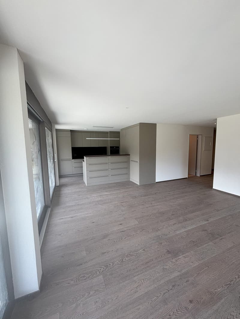 Empty room with wooden floor, white walls, kitchen with cabinets, microwave, oven, and a sliding glass door.