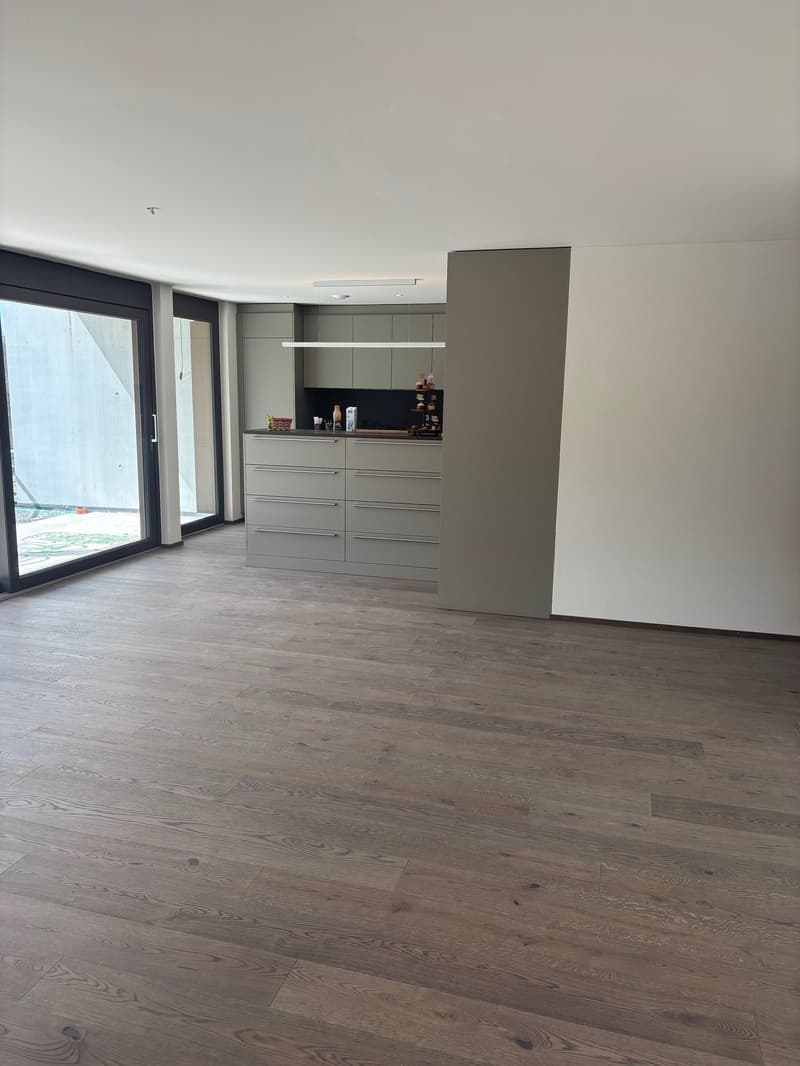 Empty room with wooden floor, white ceiling, large glass doors, kitchen with cabinets and countertop