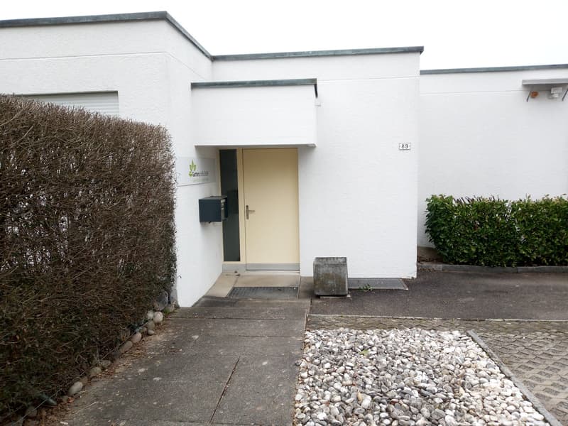 White building, entrance with a door, mailbox, gravel pathway, bushes, number 89