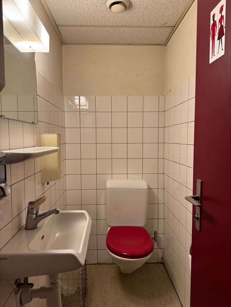 A small restroom with white tiles, a white sink, a red toilet, and a red door.