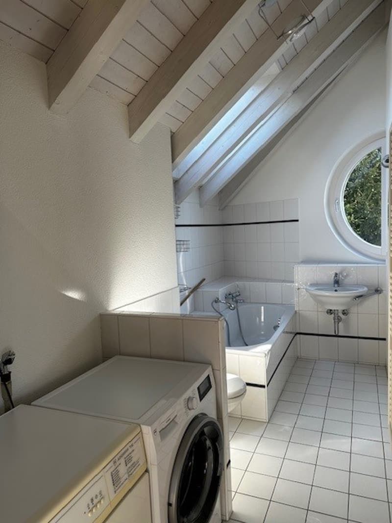 a bathroom with white tiles, a bathtub, a toilet, a washing machine, a dishwasher, and a circular window