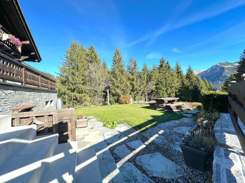 A scenic house with a view of the mountains, featuring a large green lawn, wooden picnic tables, a small patio, and a gravel pathway leading to a wooden fence.