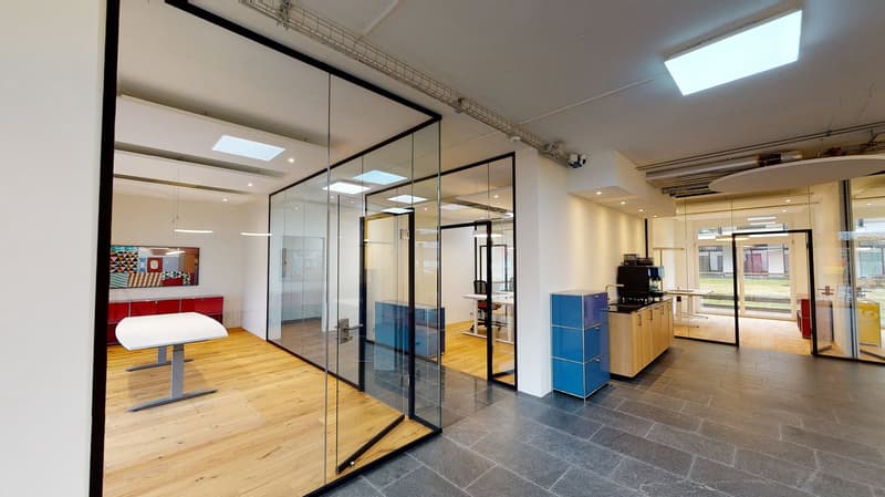 Open glass walls, wooden floors, bright white lights on ceiling, open office area with tables and chairs, blue cabinets, modern design