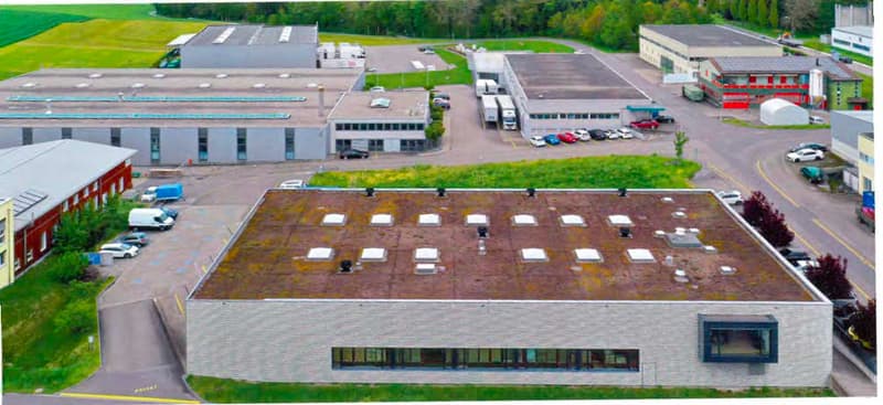 This image shows a large industrial or commercial building complex with multiple structures and a large parking lot. The buildings have a modern, industrial design with flat roofs and large windows. The parking lot in the foreground has several vehicles p