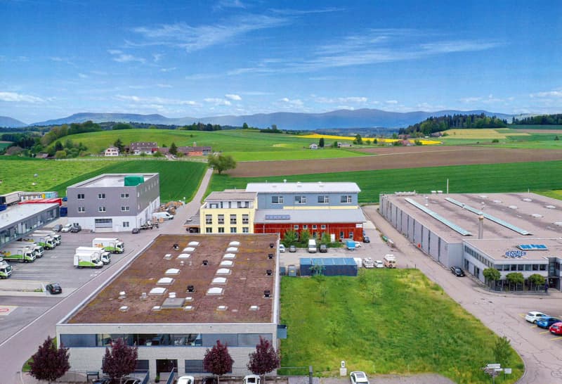 The image shows an industrial or commercial area with several multi-story buildings, warehouses, and parking lots. The surrounding landscape includes green fields, hills, and mountains in the distance, creating a scenic rural setting. There are various ve