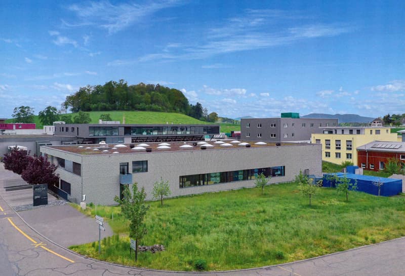 A modern, multi-story commercial building with a flat roof and large windows. The building is surrounded by a grassy area with some trees and other smaller buildings in the background. There appears to be a parking area in front of the building.