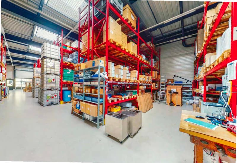 Large industrial warehouse with shelving units, storage boxes, and equipment. The warehouse has a high ceiling and is well-lit with natural light. There are various tools and supplies visible on the shelves and workbenches.