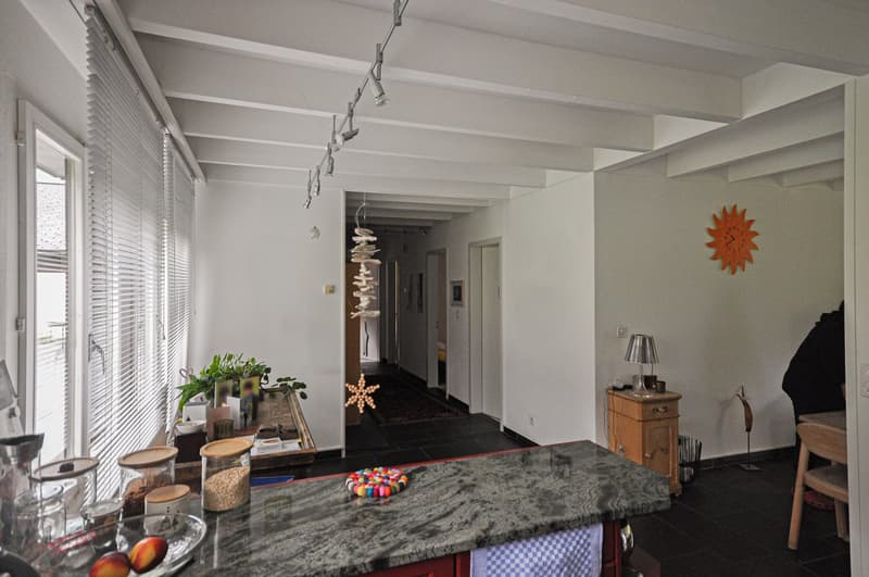 white walls, white ceiling, marble counter, kitchen island, hanging track lighting, plants, sun wall art, open hallway, lamp, wooden cabinets, dark floors