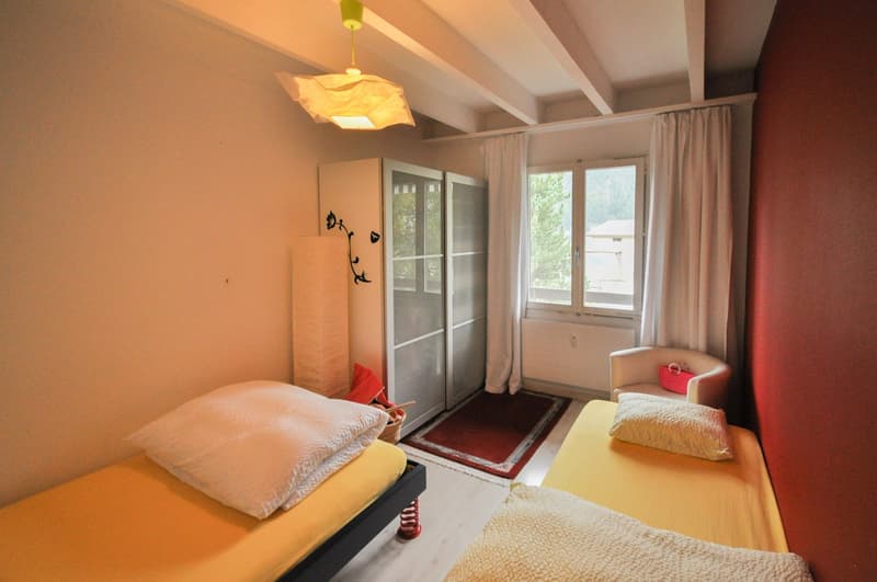 Room with two twin beds, white and yellow bedding, wooden cabinet with drawers, window with curtains, white walls, red rug in front of window, hanging lamp, chair with pink bag