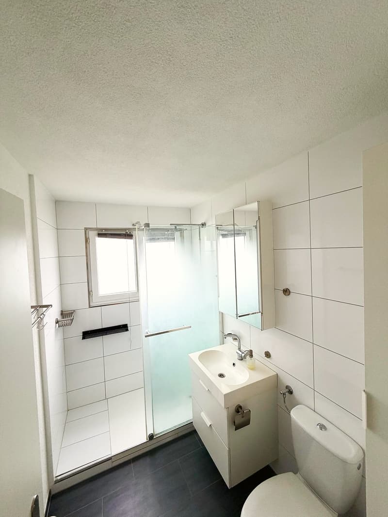Bathroom with white tiles, glass shower enclosure, white sink, mirror, toilet, and a small window