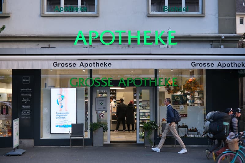 Storefront of a pharmacy with sign reading 'Apotheke', entrance, chairs, and people walking by.