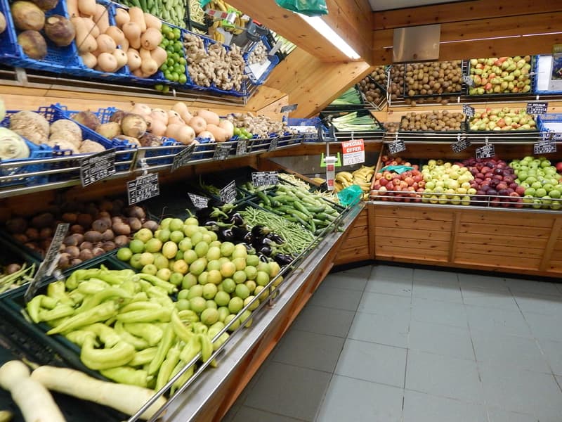 The indoor market has wooden shelves with various fruits and vegetables for sale. The shelves have blue and green plastic crates filled with different vegetables, potatoes, apples, etc. The ceiling has bright lights, and the floor is tiled.