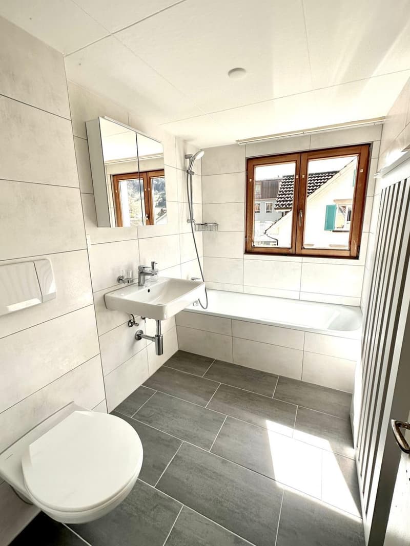 Bathroom with white walls, grey tiled floor, white bathtub, white sink, white toilet, mirror, shower head