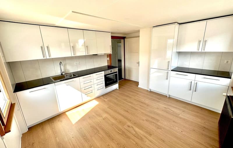 An empty kitchen with wooden floor, black countertops, white cabinetry, and a built-in oven and gas stove.