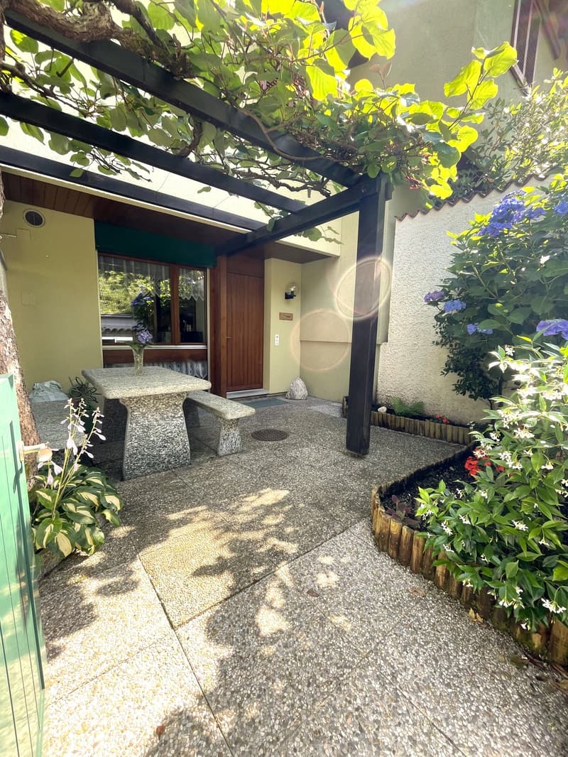 Covered patio or entryway with a wooden door, surrounded by lush greenery and flowers. The patio has a stone floor and a black metal frame overhead supporting the plants and vines. The house appears to be a single-story structure with a warm, earthy color