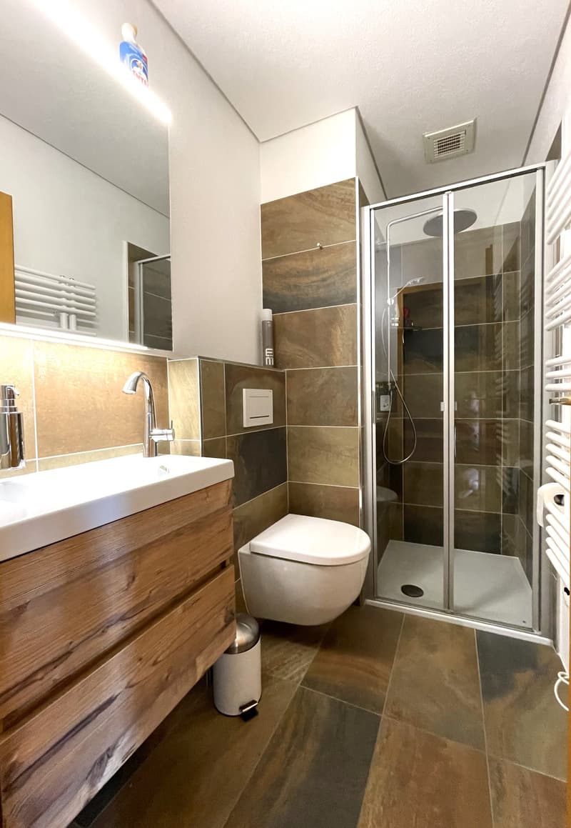 Bathroom with wooden vanity, white sink, glass shower enclosure, and tiled walls and floor
