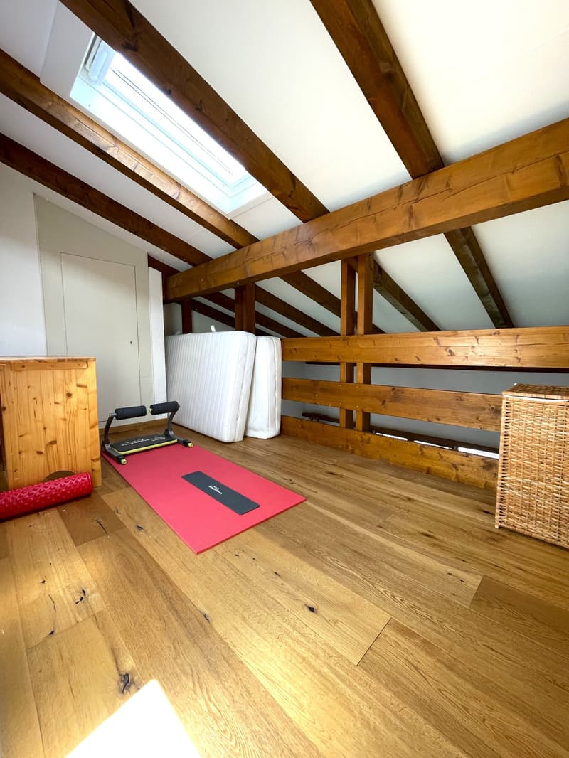 Spacious attic room with exposed wooden beams, hardwood flooring, and a skylight window. The room features a fitness mat and equipment, as well as storage space and a wicker basket.
