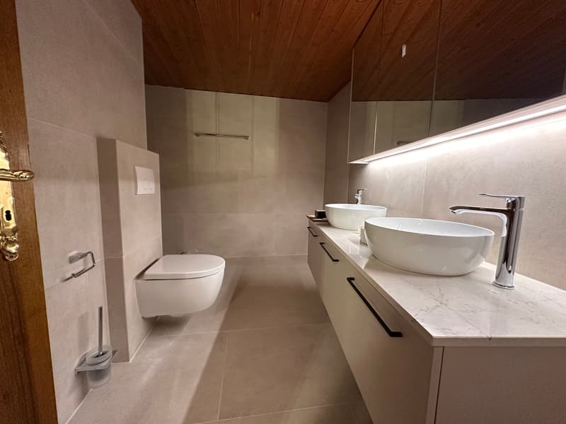 Bathroom with two sinks, white toilet, wooden ceiling, beige tiles, marble countertop, two white cabinets.