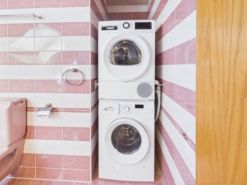 A small room with a pink and white striped wall, a toilet, and a stack of two white washing machines.