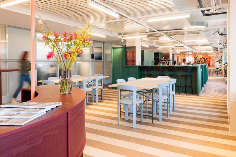 Open office space with wooden tables, chairs, and a bar area. Colorful floral arrangements and modern lighting fixtures. Bright and airy atmosphere with large windows and a casual, collaborative work environment.