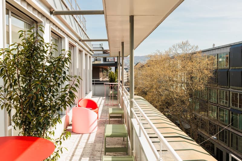 Covered balcony with seating area, potted plants, and views of surrounding buildings and trees