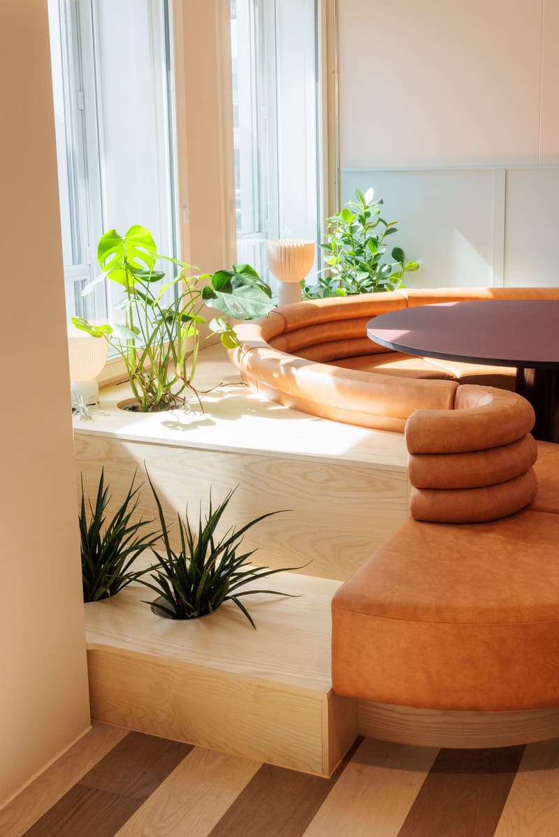 Bright and airy living room with large windows, wooden furniture, and various potted plants. The room features a round wooden table with cushioned seating, as well as a built-in bench with additional seating. The space has a warm, natural aesthetic with t