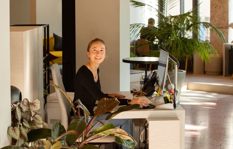 large desk with computer and phone, woman sitting, plants, glass windows