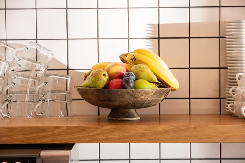 Wooden counter, glass cups, bowl of assorted fruits including bananas, apples, pears, and plums