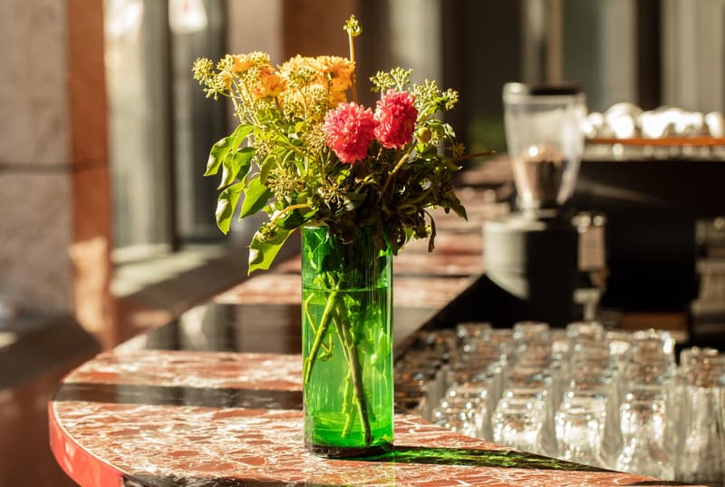 A colorful bouquet of flowers in a green glass vase, including red carnations and yellow chrysanthemums, placed on a wooden table with glassware in the background.