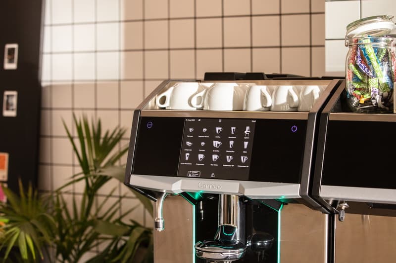 A Cameo coffee machine with a touchscreen display showing various drink options. The machine has a stainless steel body and is placed on a kitchen counter with a tiled backsplash and a potted plant nearby.
