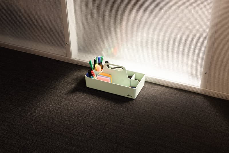 The image shows a desk organizer tray filled with various office supplies such as pens, markers, and sticky notes, placed on a dark carpeted floor near a window with a textured blind.